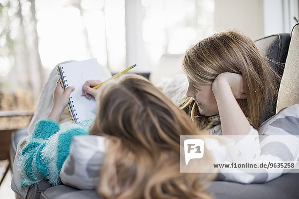 Two girls lying on a sofa  one writing into a notebook with a pencil.