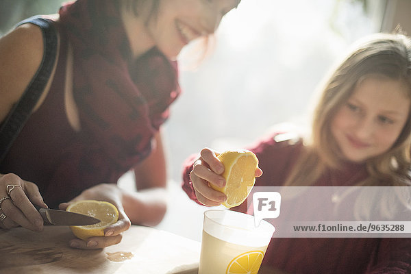 A woman and a girl sitting at a table  girl squeezing the juice from a lemon into a glass.