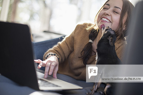 Woman lying on a sofa looking at her laptop  smiling. A small dog licking her face.