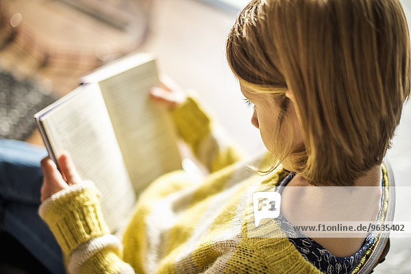High angle view of a blond girl in a yellow jumper sitting on a chair  reading a book.