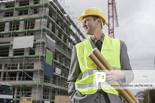 Happy male building contractor standing with blueprints and hands in pockets at a construction site