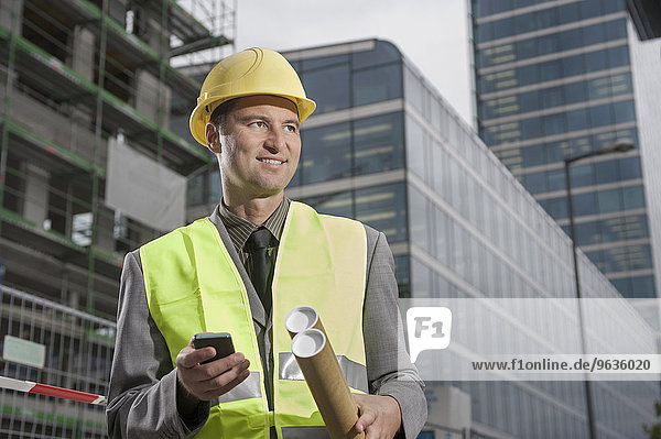 Happy site manager holding blueprints and mobile standing at a construction site