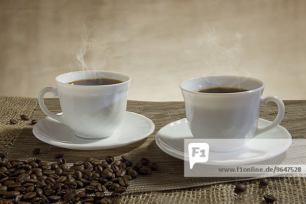 Zwei Tassen dampfender Kaffee  davor Kaffeebohnen