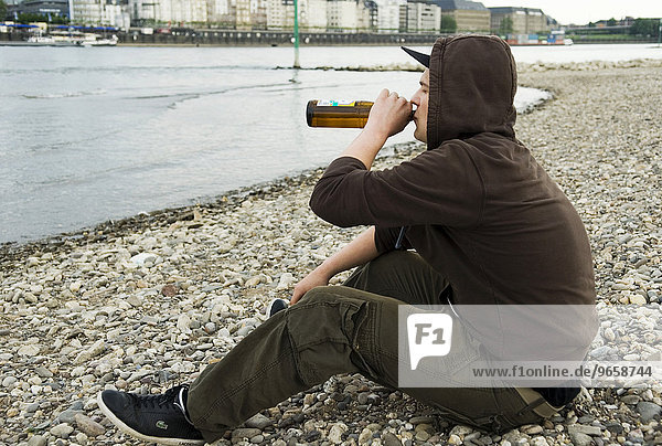 Einsamer Jugendlicher sitzt am Rheinufer und trinkt eine Flasche Bier