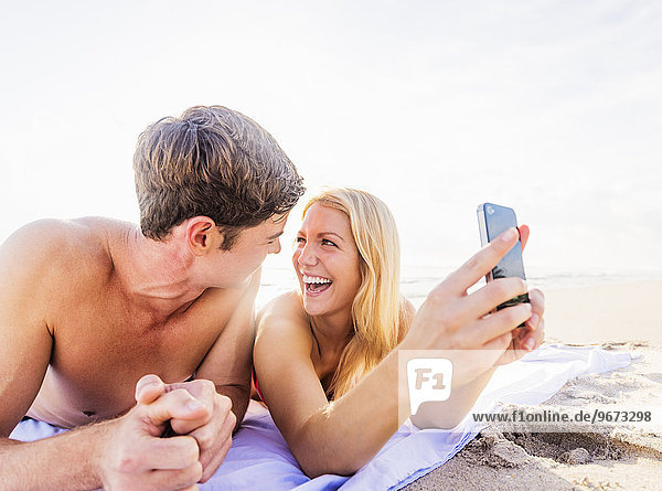 Portrait of young couple lying on blanket on beach  using smart phone