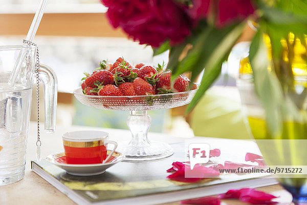 Frische Erdbeeren auf Glasschale und Mokkatasse neben verstreuten Blütenblättern auf Buch