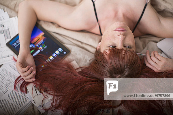 Frau auf dem Bett liegend mit digitalem Tablett und Papierkram
