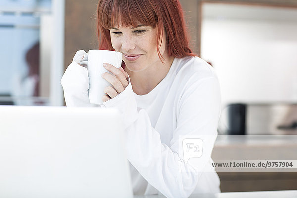 Young woman with laptop and cup of coffee in kitchen