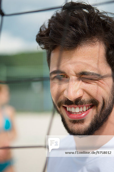 Lächelnder junger Mann funkelt am Netz des Beachvolleyballfeldes