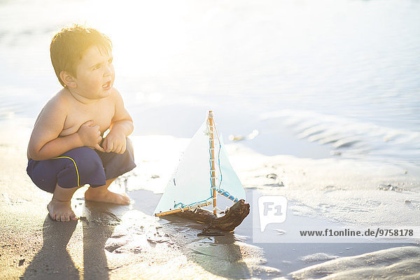 Junge am Strand spielt mit einem Spielzeug-Holzboot im Wasser