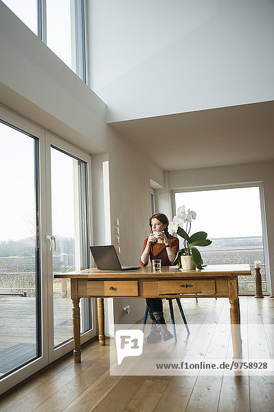 Young woman with laptop at wooden table looking out of window