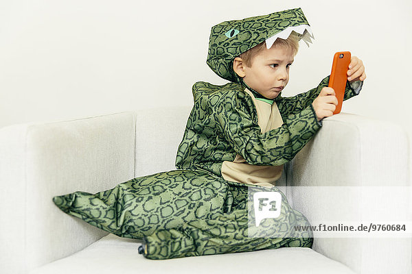 Kleiner Junge im Dinosaurierkostüm sitzend auf der Couch mit Smartphone