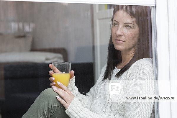 Frau mit einem Glas Orangensaft durchs Fenster schauend