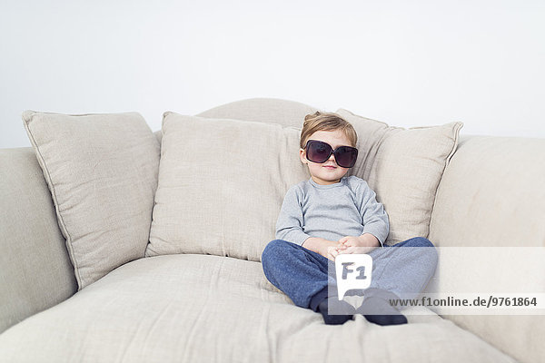 Kleiner Junge mit übergroßer Sonnenbrille auf der Couch sitzend