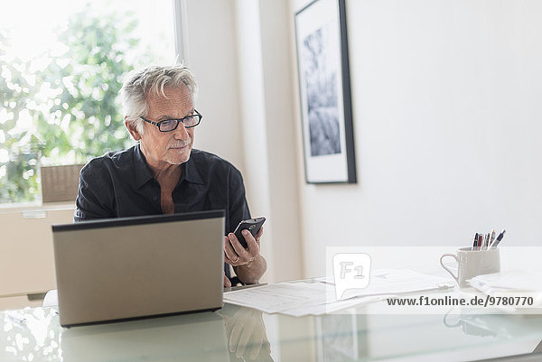 Senior man sitting in home office  using laptop and cell phone