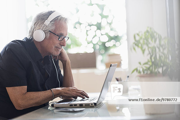 Senior man sitting in home office wearing headphones and using laptop