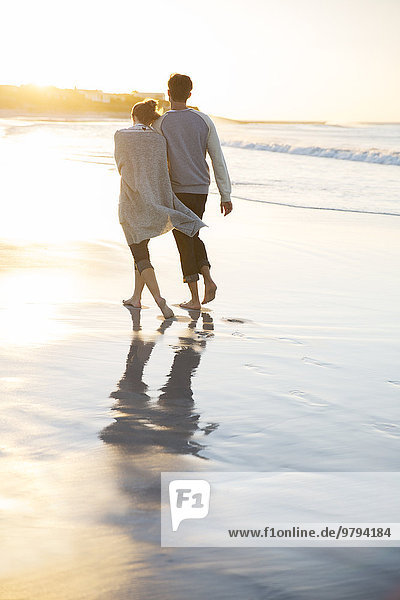 Junges Paar hält sich an den Händen und geht bei Sonnenuntergang am Strand spazieren.
