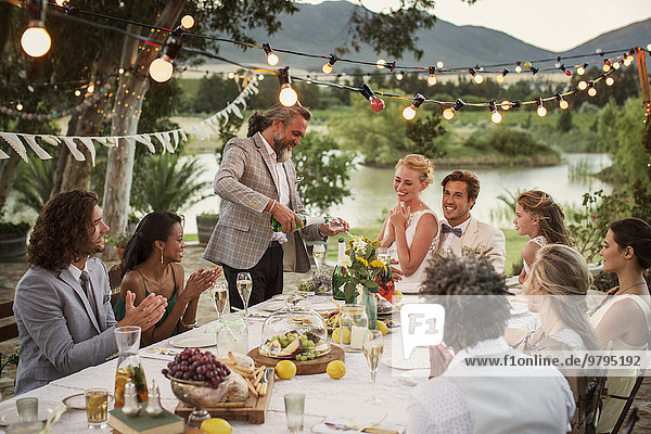 Best man pouring champagne into flutes during wedding reception in domestic garden