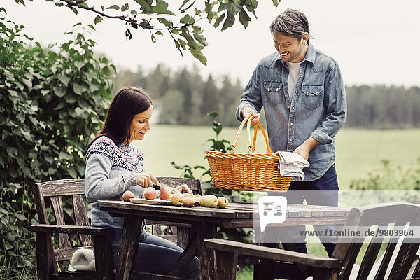 Glücklicher Mann  der den Korb auf den Tisch stellt  während die Frau auf dem Biohof Äpfel schneidet.