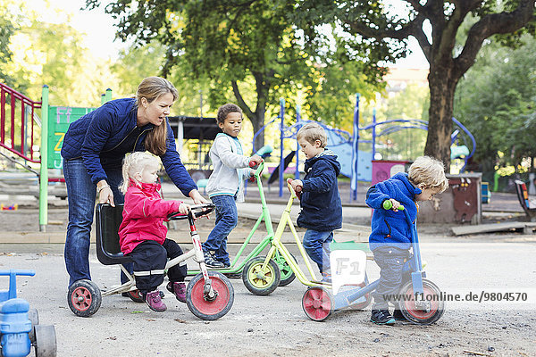 Female teacher assisting girl to ride tricycle on playground