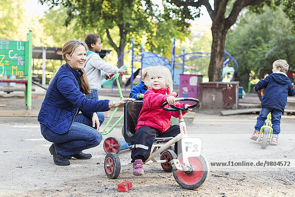 Glückliche Lehrerin  die dem Mädchen hilft  auf dem Spielplatz Dreirad zu fahren.
