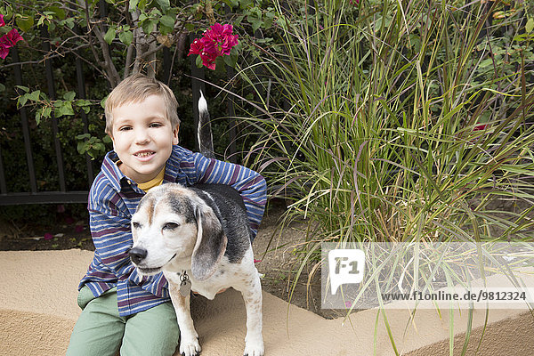 Porträt eines Jungen,  der auf einer Parkwand sitzt und den Hund umarmt.