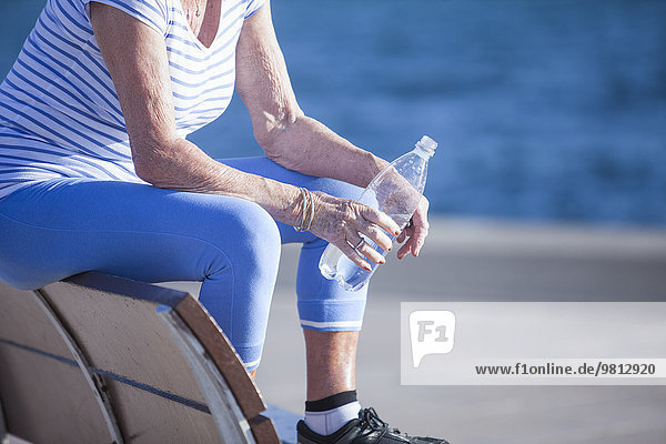 Seniorin sitzt auf einer Bank am Meer und hält Wasser in Flaschen.