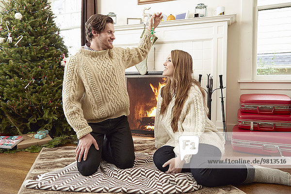 Young couple sitting on living room rug holding up christmas decoration