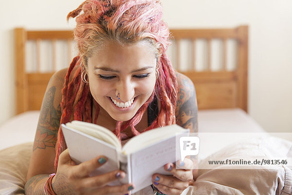 Young woman with pink dreadlocks lying on bed reading book