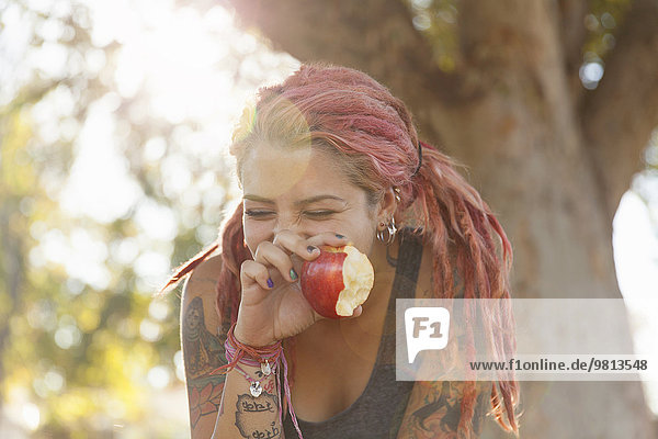 Junge Frau mit rosa Dreadlocks kichert beim Apfelessen im Park