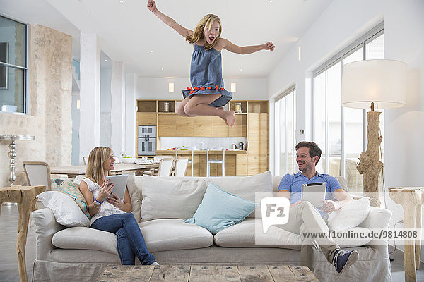 Girl jumping mid air from living room sofa whilst parents use digital tablet