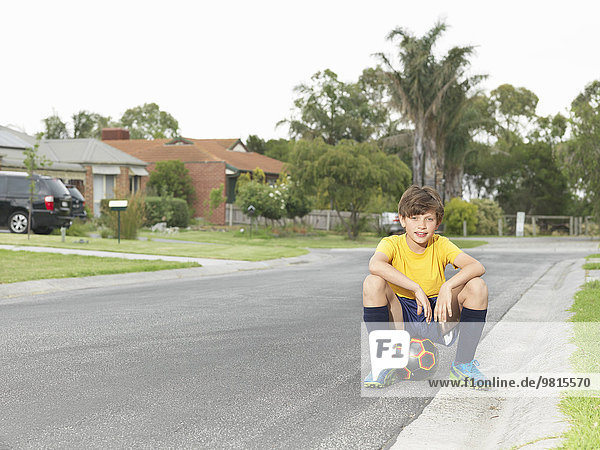 Portrait of boy sitting on soccer ball on suburban road