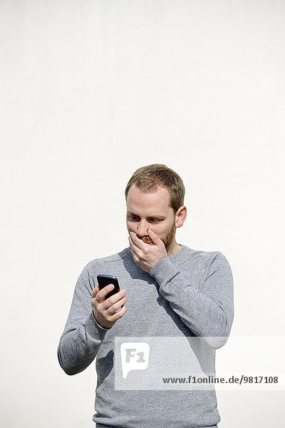 Porträt eines schockierten jungen Mannes  der sein Smartphone vor weißem Hintergrund ansieht