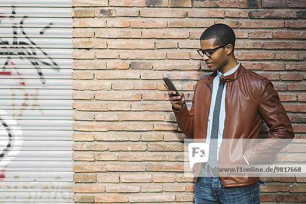 Geschäftsmann mit Smartphone in Lederjacke und Brille vor der Ziegelwand