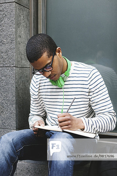Junger Mann mit grünen Kopfhörern sitzt draußen und schreibt etwas in sein Notizbuch.
