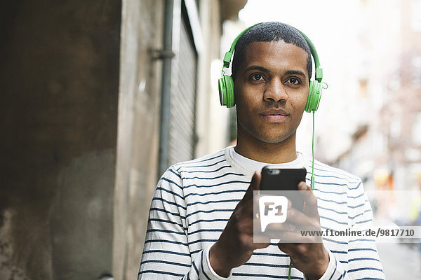 Spanien  Barcelona  Portrait eines lächelnden jungen Mannes  der Musik mit grünen Kopfhörern auf der Straße hört.