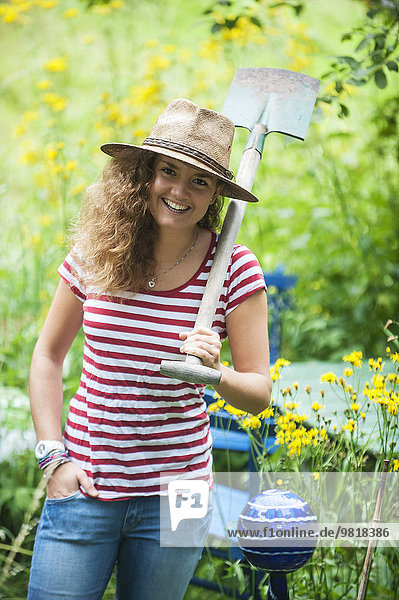 Lächelnde junge Frau mit Strohhut und Spaten im Garten