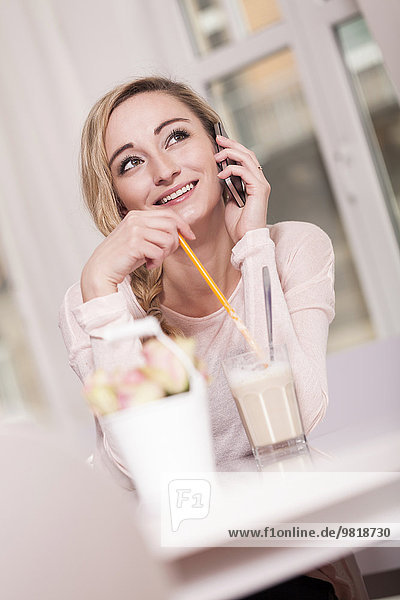 Lächelnde blonde Frau sitzt in einem Café mit einem Glas Latte Macchiato am Telefon.