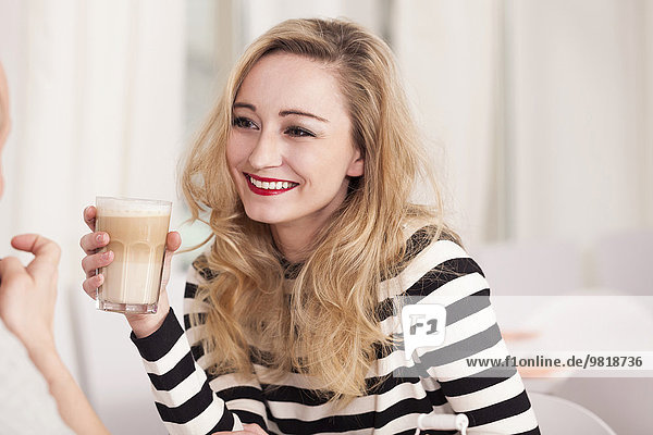 Lächelnde blonde Frau sitzt in einem Kaffee und hält ein Glas Latte Macchiato.