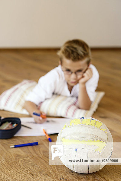 Junge macht Hausaufgaben auf Holzboden mit im Vordergrund liegendem Fußball