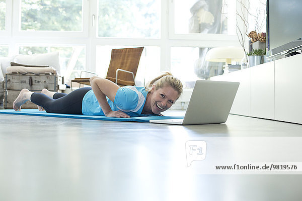 Woman with laptop exercising on gym mat in living room