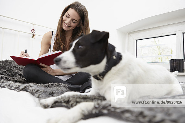 Young woman sitting on bed with dog writing diary