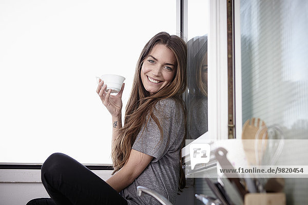 Young woman in kitchen sitting at window drinking coffee