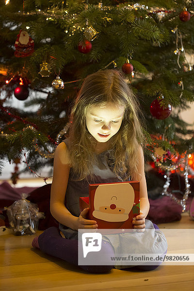 Kleines Mädchen sitzt vor dem Weihnachtsbaum und sieht sich ein Weihnachtsgeschenk an.