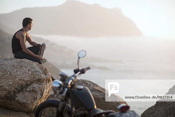 South Africa  Cape Town  motorcyclist sitting on rock at the coast enjoying view