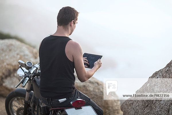 Südafrika  Kapstadt  Motorradfahrer an der Küste beim Fotografieren mit digitalem Tablett