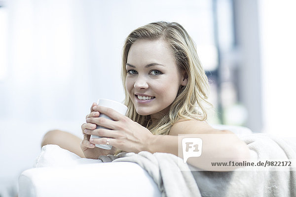 Porträt einer blonden Frau  die auf einer Couch mit einer Tasse Kaffee sitzt.