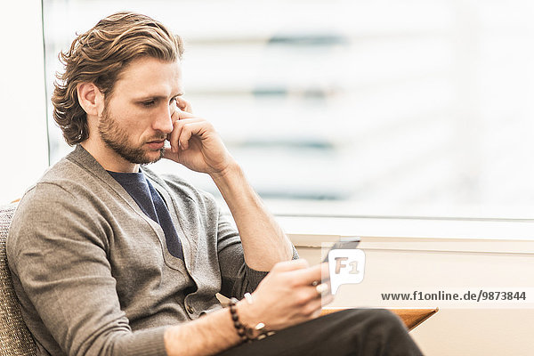 A bearded man sitting frowning and checking his phone.