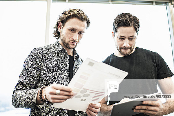 Two men standing in an office  looking at a page of printing  and referring to a digital tablet.