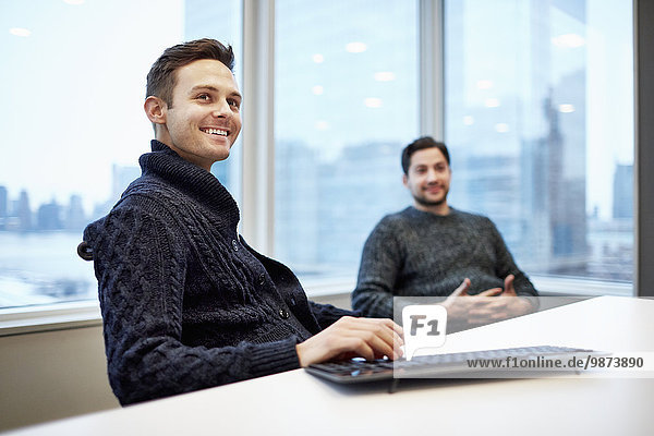 Zwei Männer in einem Büro  die an einem Schreibtisch sitzen und sich unterhalten und lächeln.